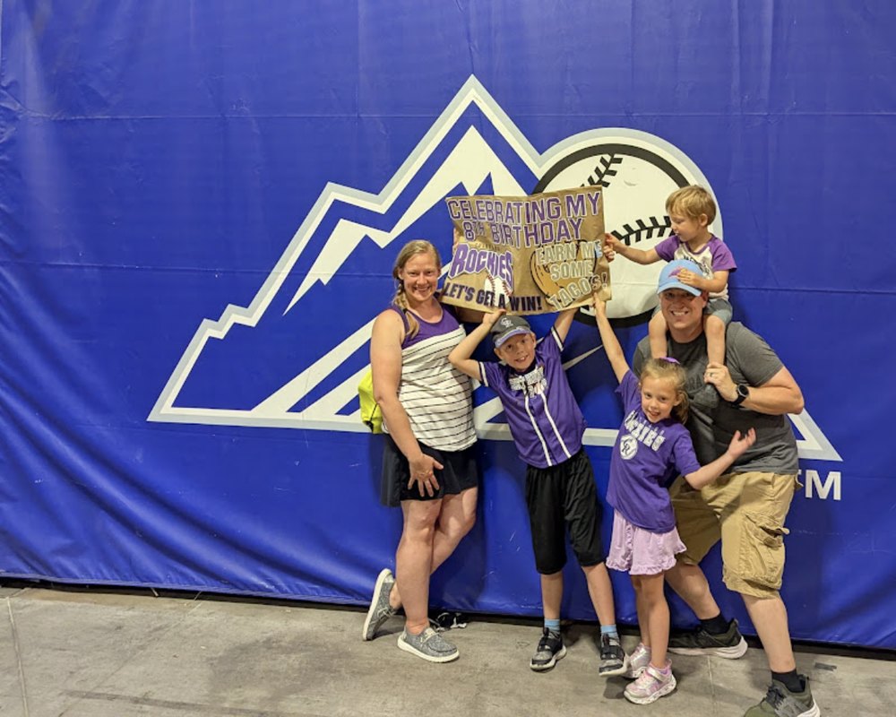 Kristen and her family at a Rockies game holding a handmade sign that says Celebrating My 8th Birthday