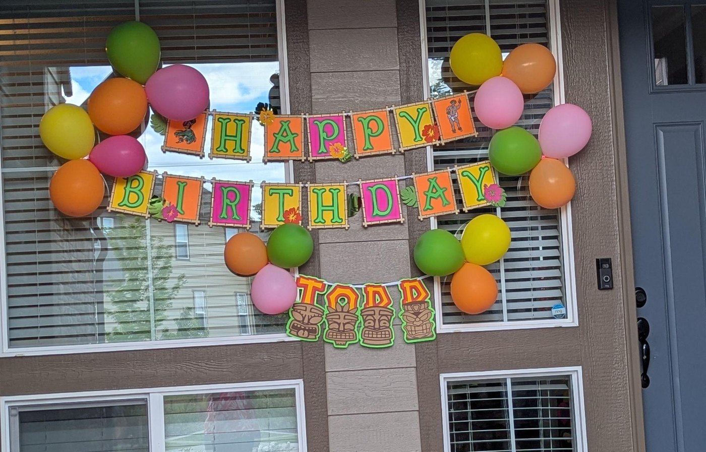A Luau Happy Birthday banner strung across a front window with balloons, and a name banner spelling TODD on tiki flags