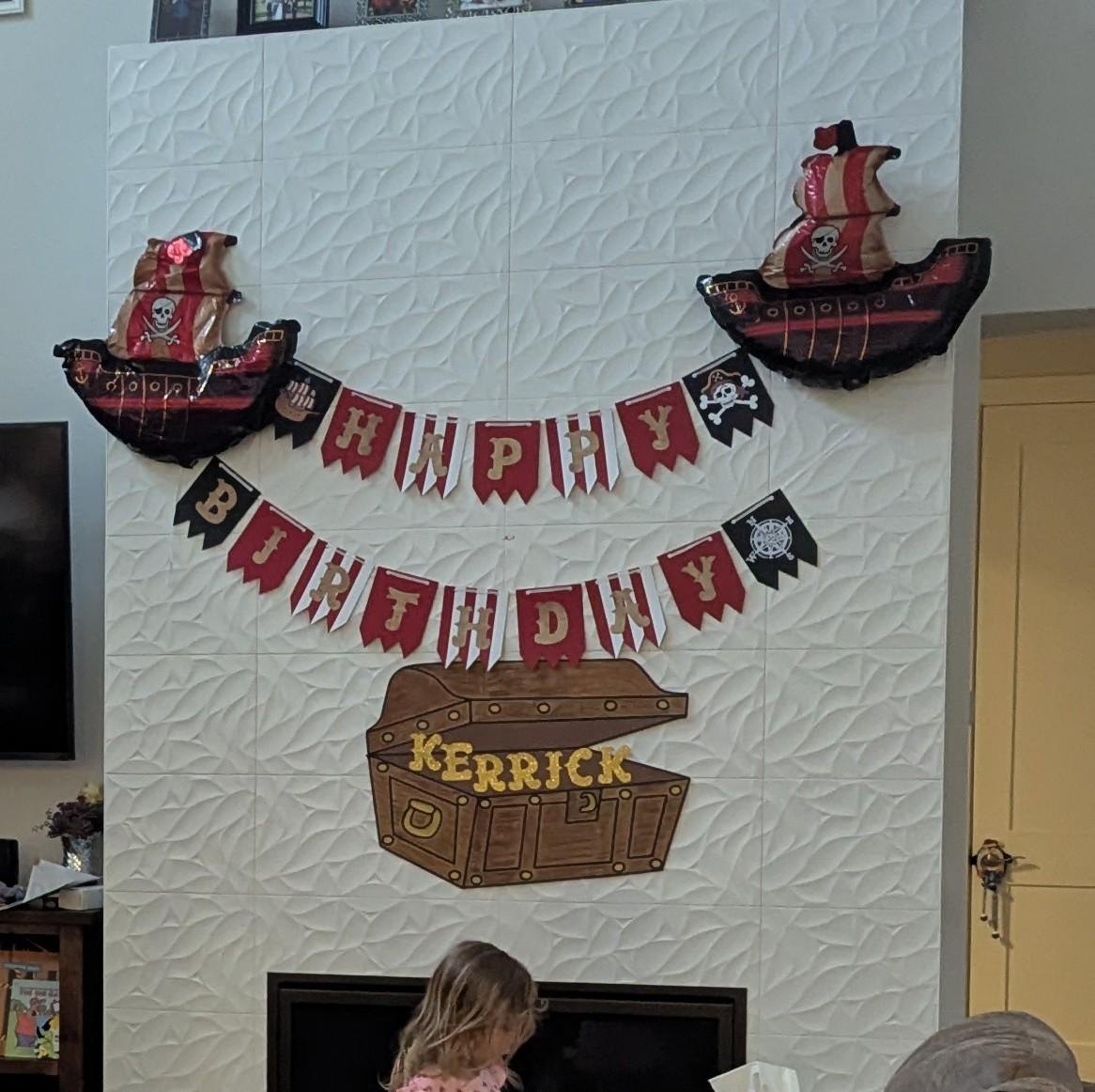A pirate Happy Birthday banner on a fireplace wall with pirate ship balloons and a treasure chest sign spelling KERRICK